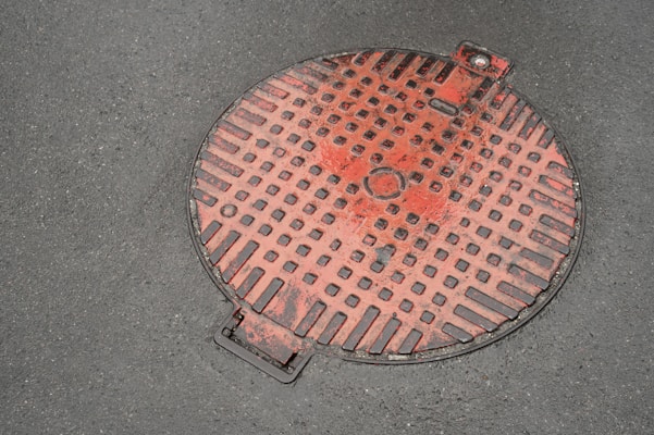 A red manhole cover with a grid pattern lies on a concrete surface. The cover has square indents and a small circular feature at the center. Some wear and chipping are visible, giving it a weathered appearance. It’s positioned slightly off-center with a metal handle visible at the edge.