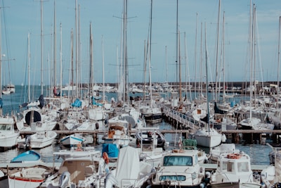 A busy marina filled with numerous sailboats and yachts tied alongside wooden docks. Masts rise high into the clear blue sky, creating a forest of poles above the boats. The harbor is bustling with activity, and the water is calm, reflecting some of the boats and the sky.