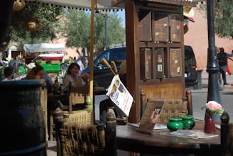 An outdoor cafe with woven chairs and tables set under a canopy. There are menus displayed on a wooden board. In the background, people are sitting and engaging in conversation. A carriage with a decorated canopy is visible, along with a parked car and some pedestrians.
