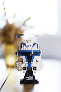 A LEGO model depicting a Star Wars character helmet, specifically Captain Rex, is placed on a wooden surface. The helmet features a striking design with predominantly white and blue bricks, detailed edges, and a display stand with a label. The background is softly blurred, giving prominence to the LEGO figure.