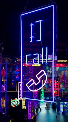 A vibrant display of neon signs with various symbols and text, predominantly featuring a large blue sign with the word 'call' and a telephone icon. The background includes multiple other neon lights in different colors like pink, yellow, and cyan, creating a lively, colorful scene.