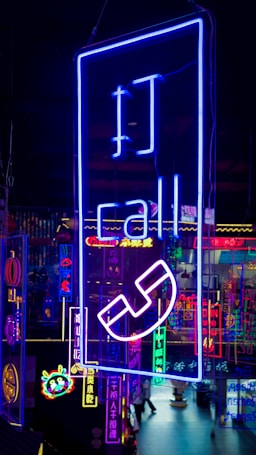 A vibrant display of neon signs with various symbols and text, predominantly featuring a large blue sign with the word 'call' and a telephone icon. The background includes multiple other neon lights in different colors like pink, yellow, and cyan, creating a lively, colorful scene.