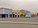A series of commercial buildings with various store signs in Arabic and English, including automotive parts companies. The buildings are mostly closed with shutters down, and there is a truck and a parked white car in front. The scene is empty with an overcast sky.