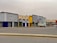 A series of commercial buildings with various store signs in Arabic and English, including automotive parts companies. The buildings are mostly closed with shutters down, and there is a truck and a parked white car in front. The scene is empty with an overcast sky.