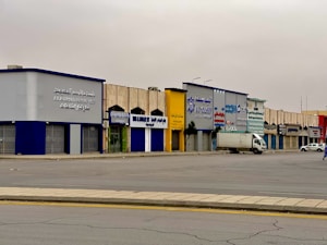 A series of commercial buildings with various store signs in Arabic and English, including automotive parts companies. The buildings are mostly closed with shutters down, and there is a truck and a parked white car in front. The scene is empty with an overcast sky.