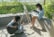 Two individuals are outdoors in a sunny setting surrounded by greenery. One person is seated on the ground playing a black acoustic guitar, while the other is seated on a concrete bench, painting on an easel. The scene is bathed in natural light, casting soft shadows.