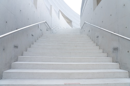 A set of wide, concrete stairs is flanked by steel railings on both sides. The gray concrete walls on either side are smooth with evenly spaced embedded fixtures. The staircase leads upwards towards a bright area, indicating an outdoor or well-lit space at the top.