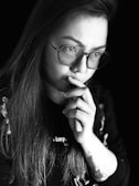 A person with long hair and glasses is deep in thought. They are holding their hand to their mouth and appear to be contemplating something. The image is in black and white, adding a dramatic and introspective feel.