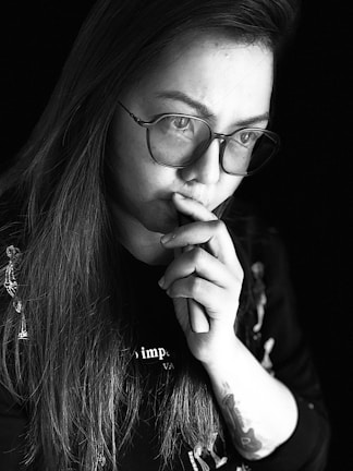 A person with long hair and glasses is deep in thought. They are holding their hand to their mouth and appear to be contemplating something. The image is in black and white, adding a dramatic and introspective feel.