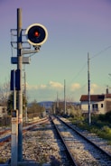 A railway signal with a red light is positioned beside a set of train tracks that stretch into the distance, flanked by fencing and utility poles. In the background, houses and trees are visible under a clear sky with scattered clouds and distant hills.
