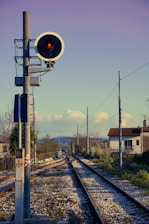 A railway signal with a red light is positioned beside a set of train tracks that stretch into the distance, flanked by fencing and utility poles. In the background, houses and trees are visible under a clear sky with scattered clouds and distant hills.