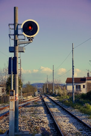 A railway signal with a red light is positioned beside a set of train tracks that stretch into the distance, flanked by fencing and utility poles. In the background, houses and trees are visible under a clear sky with scattered clouds and distant hills.