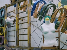 A weathered playground area featuring colorful tires stacked vertically, a mannequin torso with a smiley face and a missing head resting on a chair, and a wooden climbing frame. Various playground equipment is visible in the background, with some paint peeling and signs of wear.