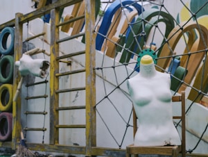 A weathered playground area featuring colorful tires stacked vertically, a mannequin torso with a smiley face and a missing head resting on a chair, and a wooden climbing frame. Various playground equipment is visible in the background, with some paint peeling and signs of wear.