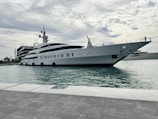 A large luxury yacht is docked at a harbor with a modern building and clear skies in the background. The yacht is expansive and sleek, with a white exterior and multiple decks. People are seen on the deck, indicating activity.