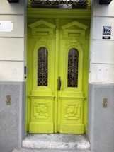 A vibrant lime-green double door featuring intricate woodwork and glass panels with ornate ironwork designs. The surrounding wall is painted in a light, neutral color with decorative elements, and there are doorbell buttons on either side. The number 12 appears in blue and white on a plaque next to the door.