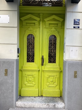 A vibrant lime-green double door featuring intricate woodwork and glass panels with ornate ironwork designs. The surrounding wall is painted in a light, neutral color with decorative elements, and there are doorbell buttons on either side. The number 12 appears in blue and white on a plaque next to the door.