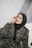 A person posing in a patterned outfit with a black hijab, wearing sunglasses on top of their head. The background consists of draped fabric, and the individual is seated with an arm resting on a white surface.