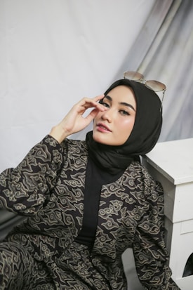 A person posing in a patterned outfit with a black hijab, wearing sunglasses on top of their head. The background consists of draped fabric, and the individual is seated with an arm resting on a white surface.