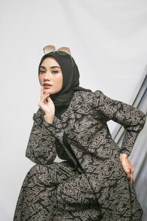 A woman wearing a black headscarf and stylish patterned outfit poses with one hand touching her chin. She is wearing sunglasses on top of her head and is positioned against a simple background with draped fabric.