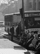 A city street scene with a bus stop where several individuals are boarding a bus. Motor scooters are parked nearby, and there is a mixture of light and shadow due to the time of day, possibly morning or afternoon. The atmosphere appears busy, with urban life in motion.