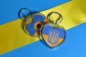 Two heart-shaped keychains are placed on a surface with a design featuring horizontal stripes of blue and yellow. One keychain displays a sunflower graphic with green leaves, and the other has a trident emblem. The background is composed of vivid blue and yellow colors.