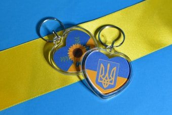 Two heart-shaped keychains are placed on a surface with a design featuring horizontal stripes of blue and yellow. One keychain displays a sunflower graphic with green leaves, and the other has a trident emblem. The background is composed of vivid blue and yellow colors.