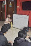 A woman wearing a headscarf and mask is engaged in an interactive session with two young people. She is pointing at a whiteboard filled with comparative adjectives for characters in a list. The setting appears to be an indoor room with red walls, a tiled floor, and closed wooden doors.