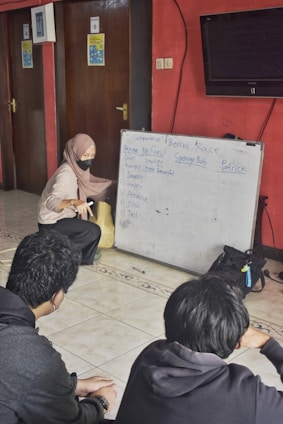 A woman wearing a headscarf and mask is engaged in an interactive session with two young people. She is pointing at a whiteboard filled with comparative adjectives for characters in a list. The setting appears to be an indoor room with red walls, a tiled floor, and closed wooden doors.