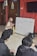 A woman wearing a headscarf and mask is engaged in an interactive session with two young people. She is pointing at a whiteboard filled with comparative adjectives for characters in a list. The setting appears to be an indoor room with red walls, a tiled floor, and closed wooden doors.