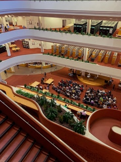 A multi-level interior space with multiple floors visible, each lined with bookshelves and people engaging in various activities. The ground floor is hosting an event where a sizable audience is seated facing a speaker. The space features red carpeting and an abundance of greenery along the balconies.
