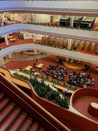 A multi-level interior space with multiple floors visible, each lined with bookshelves and people engaging in various activities. The ground floor is hosting an event where a sizable audience is seated facing a speaker. The space features red carpeting and an abundance of greenery along the balconies.