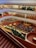 A multi-level interior space with multiple floors visible, each lined with bookshelves and people engaging in various activities. The ground floor is hosting an event where a sizable audience is seated facing a speaker. The space features red carpeting and an abundance of greenery along the balconies.