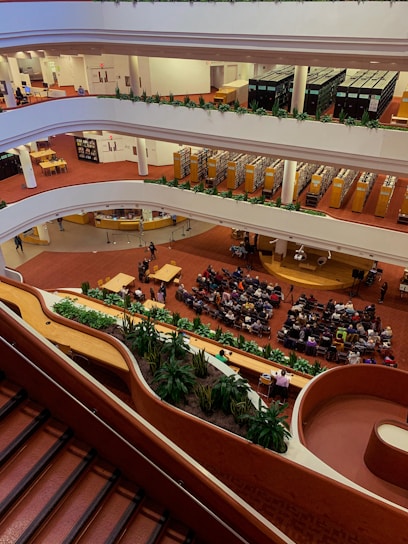 A multi-level interior space with multiple floors visible, each lined with bookshelves and people engaging in various activities. The ground floor is hosting an event where a sizable audience is seated facing a speaker. The space features red carpeting and an abundance of greenery along the balconies.