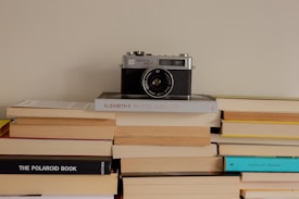 A vintage camera is placed on top of a stack of books, suggesting a nostalgic or retro theme. The books are arranged neatly, with titles visible, adding to the scholarly ambiance. The overall setting is minimalistic and aesthetically pleasing.