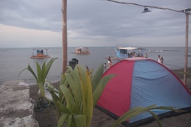 A beach setting with a large red and blue tent situated on the sand. Several people are interacting near the shore, and small huts on bamboo rafts are floating in the calm sea. Palm leaves and trunks create a tropical atmosphere. The sky is overcast, indicating an impending change in weather.