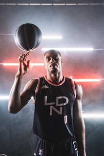 A basketball player in a black jersey with 'LDN' written on it holds a spinning basketball on one finger. The background features a modern, industrial look with horizontal neon lights in red and white, creating a dynamic atmosphere.