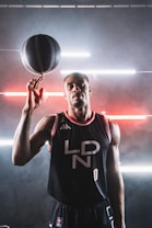 A basketball player in a black jersey with 'LDN' written on it holds a spinning basketball on one finger. The background features a modern, industrial look with horizontal neon lights in red and white, creating a dynamic atmosphere.