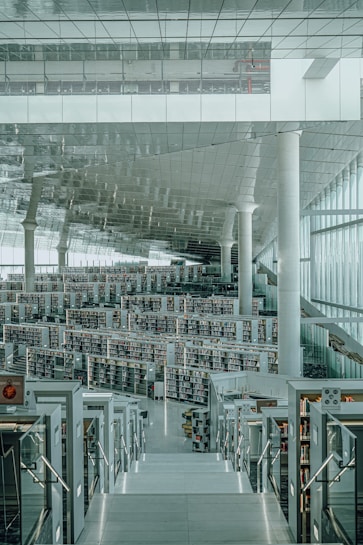 A modern library with a vast open space, featuring multiple levels and rows of bookshelves. The architecture includes sleek, reflective surfaces and large windows allowing natural light to flood the interior. White columns and a mirrored ceiling add to the spacious and futuristic feel of the library.