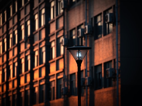 A modern streetlamp stands in front of a tall brick building with multiple windows. The building is bathed in warm sunlight, creating a golden hue. The presence of air conditioning units on the building's facade indicates an urban setting.