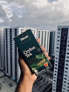 A hand holding a box of Dilmah Ceylon Tea, prominently displayed against a background of high-rise buildings under a cloudy sky. The tea box is dark green with images of tea leaves and a small cup of tea, containing details about the tea blend.