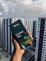 A hand holding a box of Dilmah Ceylon Tea, prominently displayed against a background of high-rise buildings under a cloudy sky. The tea box is dark green with images of tea leaves and a small cup of tea, containing details about the tea blend.