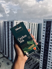 A hand holding a box of Dilmah Ceylon Tea, prominently displayed against a background of high-rise buildings under a cloudy sky. The tea box is dark green with images of tea leaves and a small cup of tea, containing details about the tea blend.