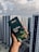 A hand holding a box of Dilmah Ceylon Tea, prominently displayed against a background of high-rise buildings under a cloudy sky. The tea box is dark green with images of tea leaves and a small cup of tea, containing details about the tea blend.