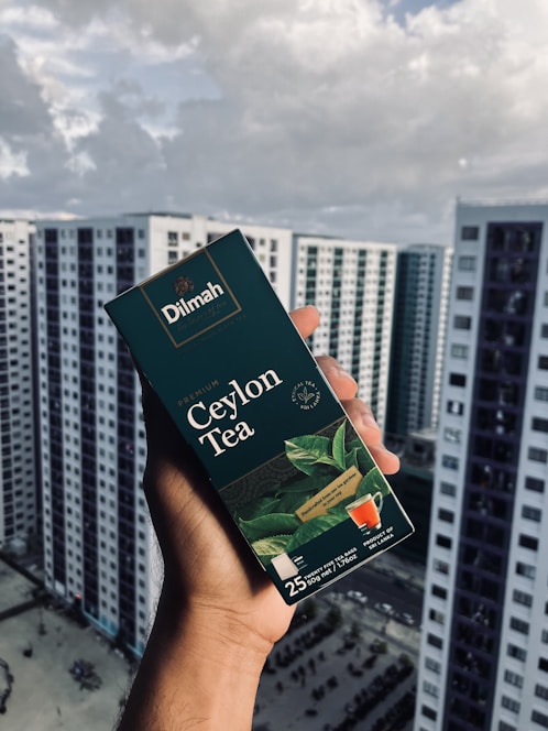 A hand holding a box of Dilmah Ceylon Tea, prominently displayed against a background of high-rise buildings under a cloudy sky. The tea box is dark green with images of tea leaves and a small cup of tea, containing details about the tea blend.