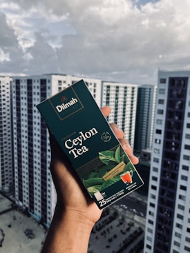 A hand holding a box of Dilmah Ceylon Tea, prominently displayed against a background of high-rise buildings under a cloudy sky. The tea box is dark green with images of tea leaves and a small cup of tea, containing details about the tea blend.