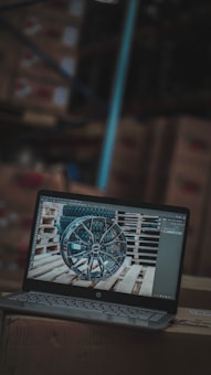 A laptop is displaying an image of a car wheel on its screen, placed on top of cardboard boxes in what appears to be a warehouse. The surroundings are blurred, with visible shelves and boxes in the background.