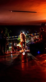 A band is performing on stage at night, with a singer in the center facing away from the camera, surrounded by band members playing instruments. The stage is illuminated with warm, orange lighting, complemented by string lights and banners in the background.