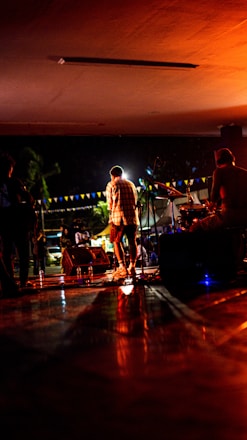 A band is performing on stage at night, with a singer in the center facing away from the camera, surrounded by band members playing instruments. The stage is illuminated with warm, orange lighting, complemented by string lights and banners in the background.