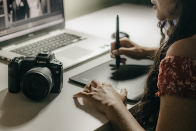 A person uses a stylus to work on a graphic tablet beside a laptop. A digital camera is also placed on the table, indicating a creative workspace. The focus is on the hand holding the stylus, illustrating an active engagement in a digital design or photo editing task.